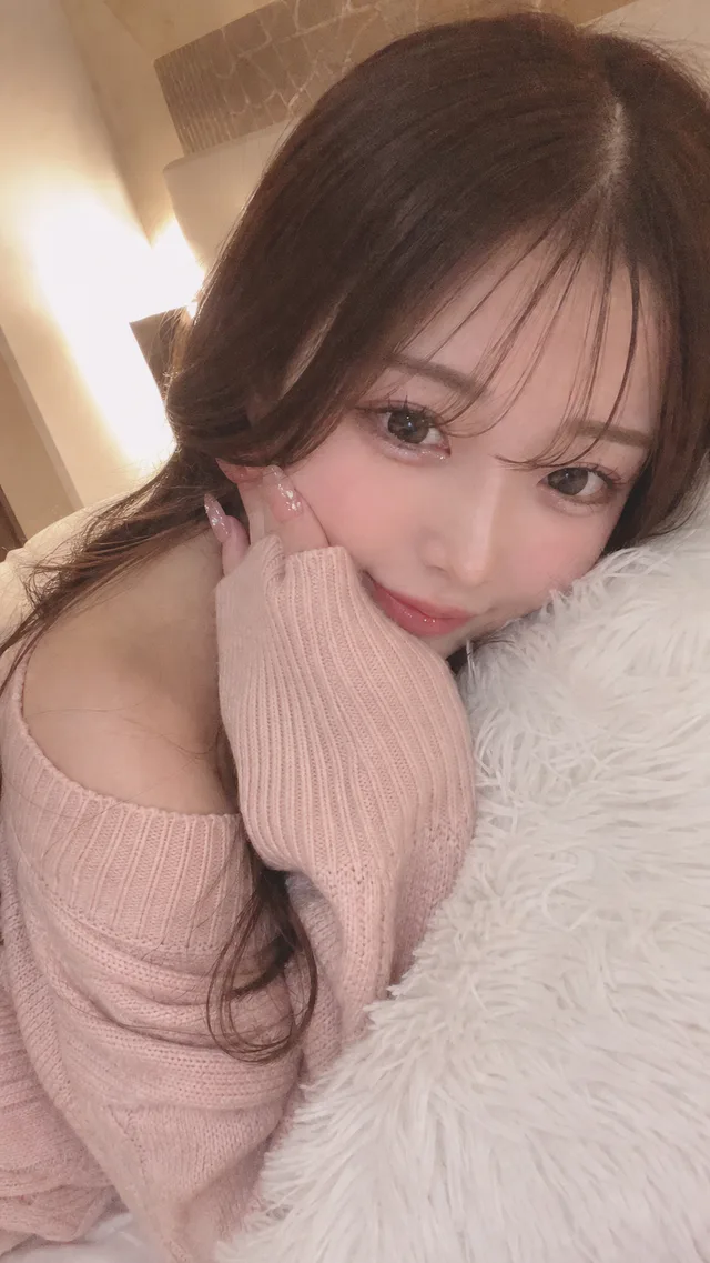 暖色の寝室で白いファークッションに顔を寄せ、淡いピンクのオフショルダーニットを着て頬に手を添える成人日本人女性のセルフィー