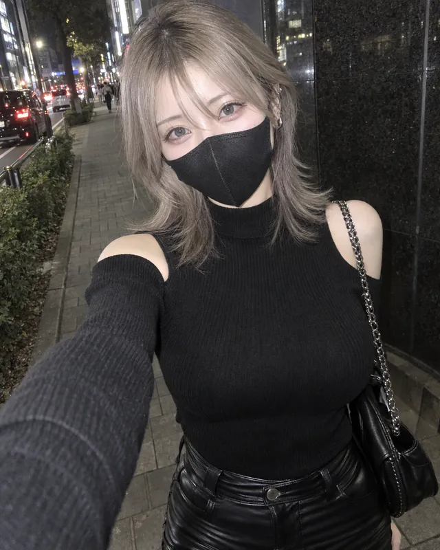 夜の街で、アッシュベージュのウルフヘアと黒マスク、肩開きニット、レザーパンツ姿の女性が自撮りしている