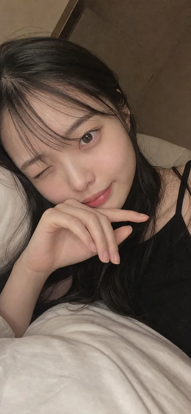 暖色の寝室で白いシーツに横になり、黒いルームウェア姿でウインクする日本人女性のスマホセルフィ