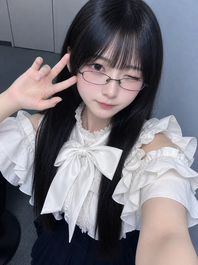青灰色の室内で白い大きなリボンと細い黒縁眼鏡を着け、片目を閉じて左手を上げる黒髪の成人女性