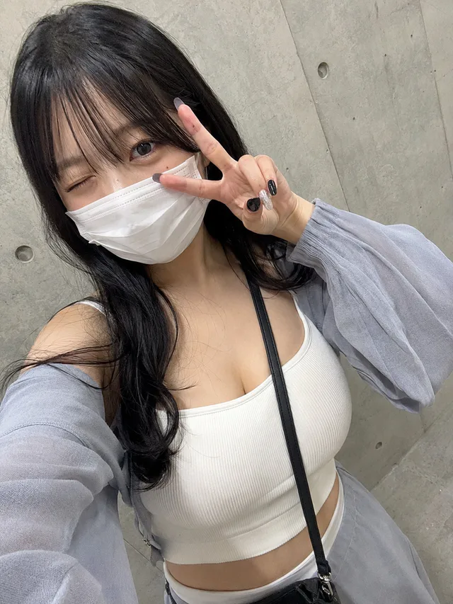 コンクリート壁の前で、白いマスクと白リブトップス、淡いグレーカーデを着た黒髪の女性がウインクしてピースサインをする自撮り
