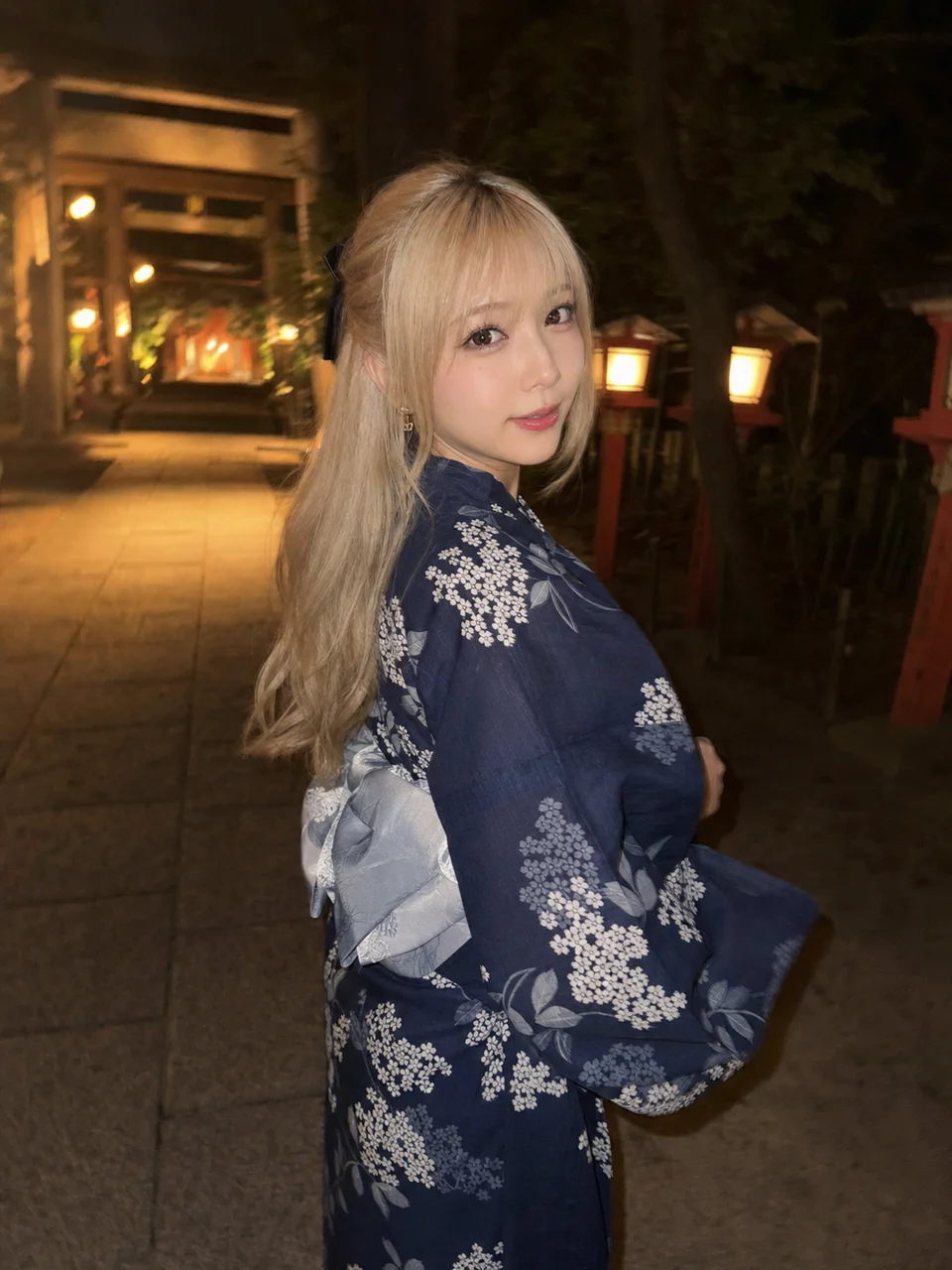 夜の神社参道で、紺色の紫陽花柄浴衣を着たプラチナブロンドの成人女性がスマホフラッシュに振り返るAI生成写真