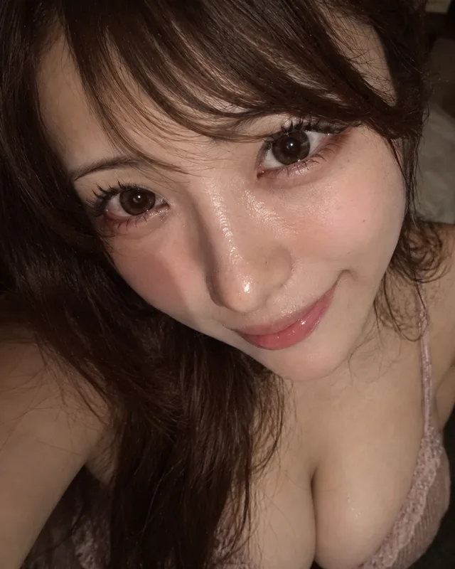 薄暗い室内でピンクのレーストップを着た成人日本人女性が、大きな瞳と艶のある肌を見せて微笑む超接近ハイアングル自撮りのAI生成画像