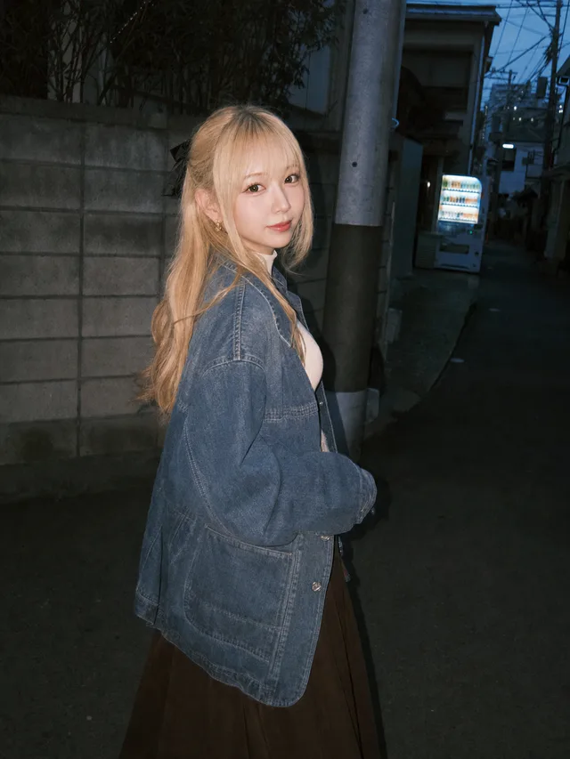 夕暮れの住宅街の路地で、デニムジャケットを羽織ったプラチナブロンドの成人女性が使い捨てカメラのフラッシュに振り向くAI生成写真