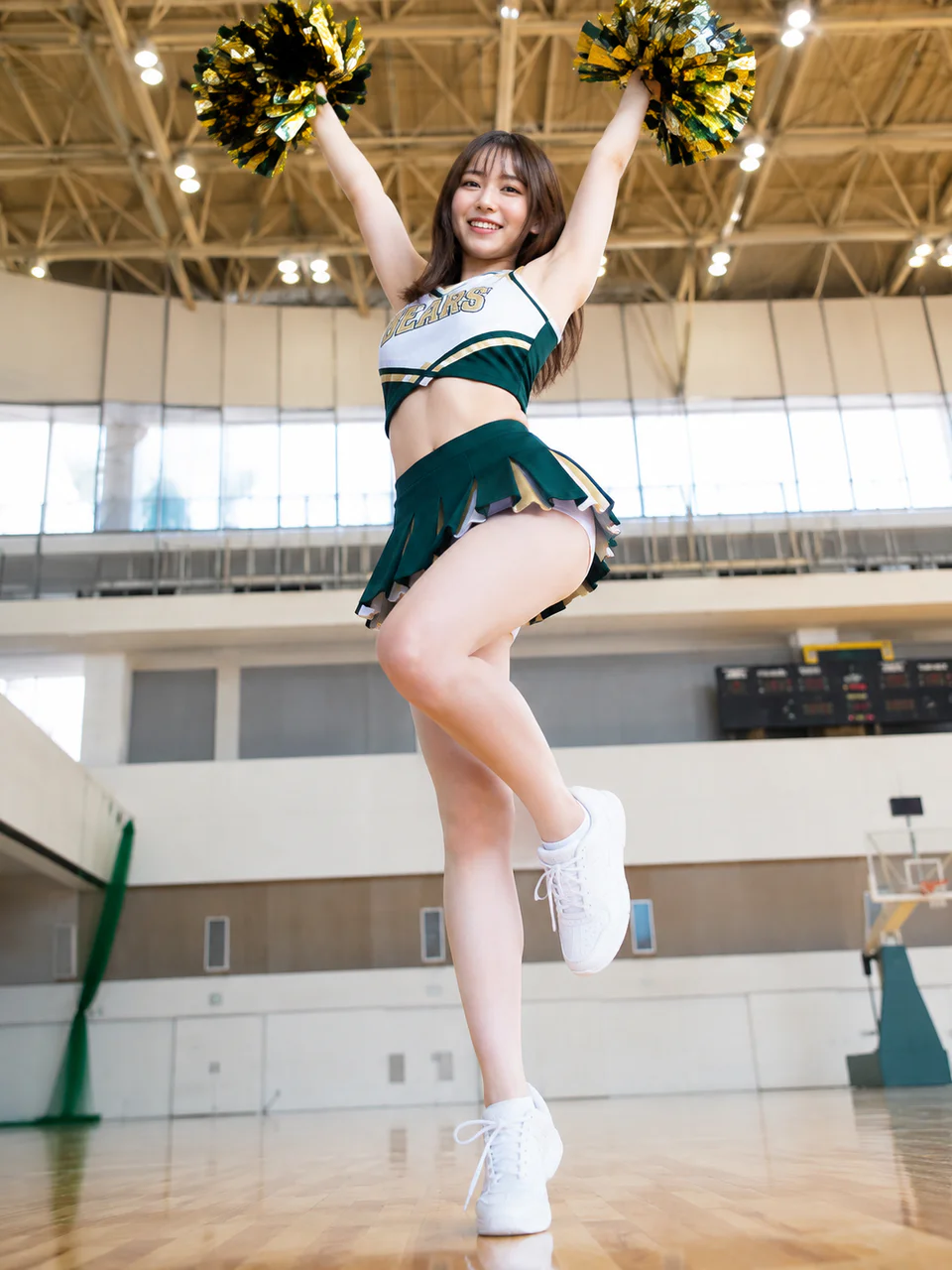 明るい体育館で金と緑のポンポンを上げ、片脚を曲げてポーズを取るチアガール。