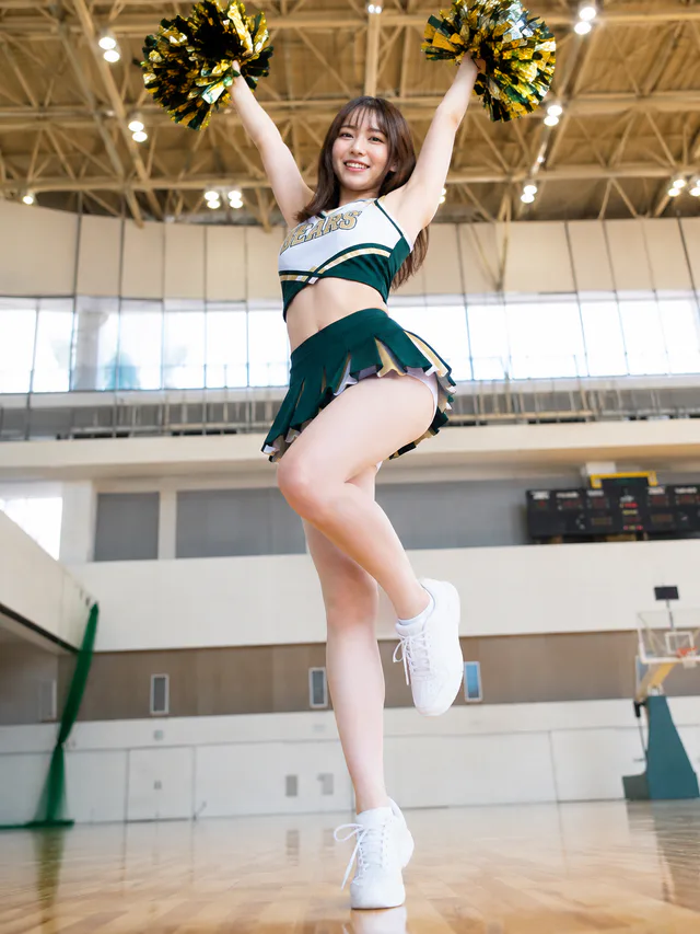 明るい体育館で金と緑のポンポンを上げ、片脚を曲げてポーズを取るチアガール。
