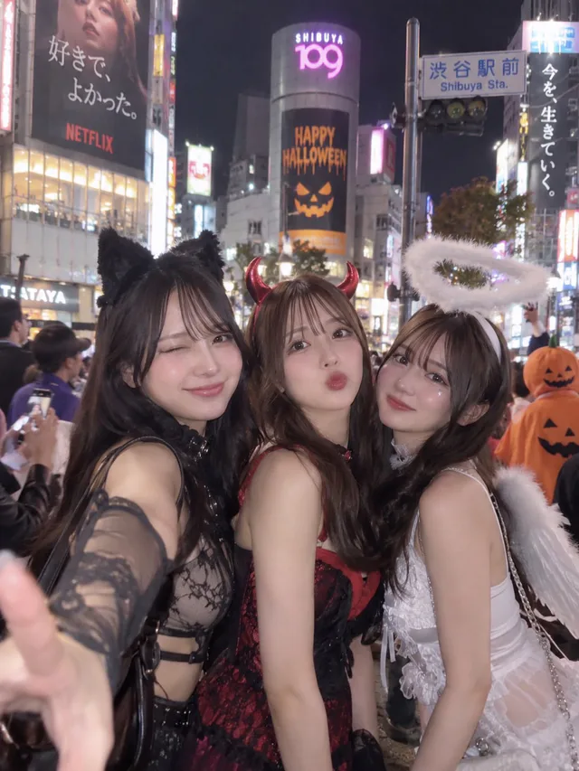 猫耳の黒い衣装、赤い悪魔衣装、白い天使衣装の3人の成人女性が、夜の渋谷で身を寄せて自撮りするAI生成画像