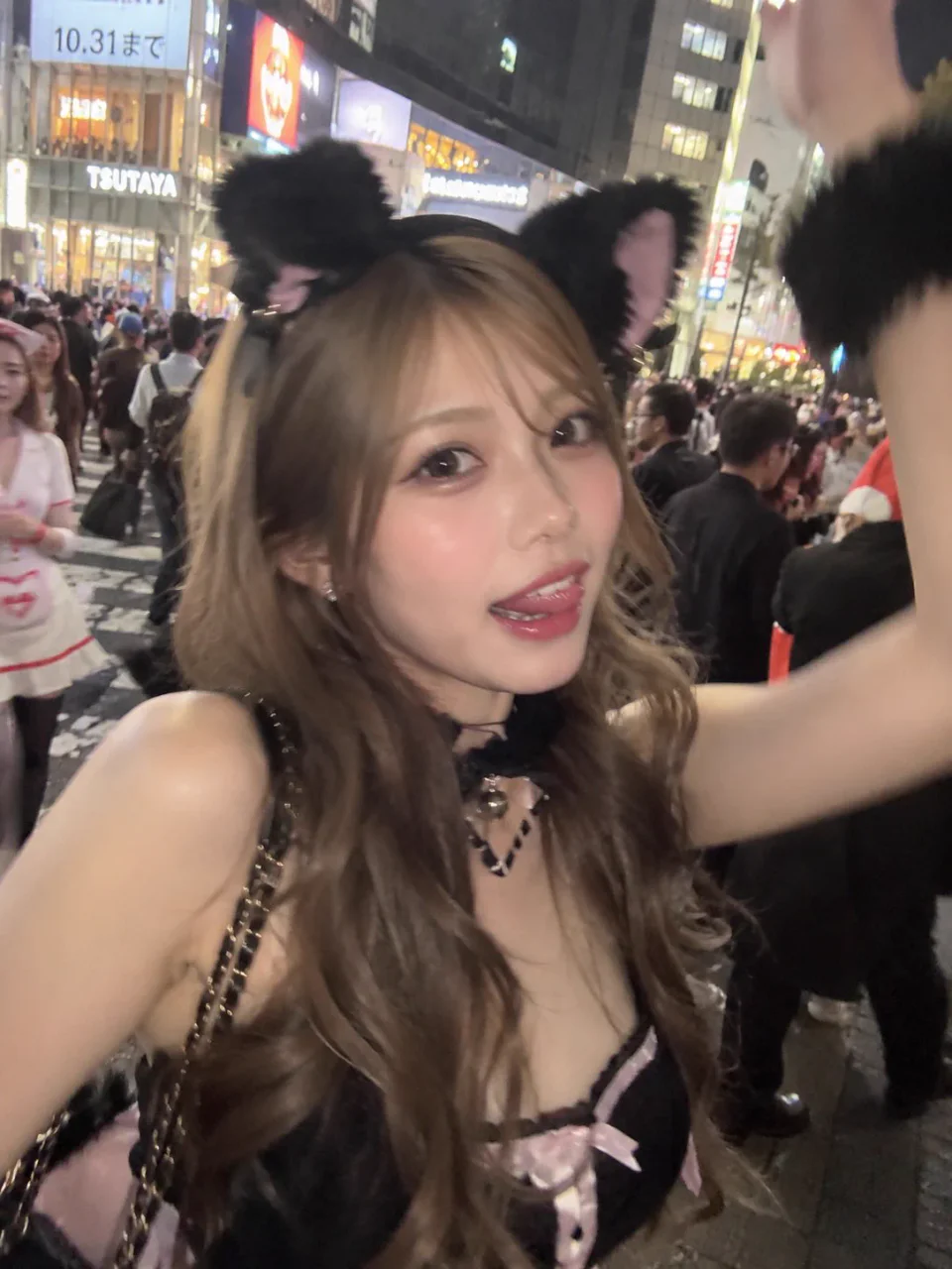夜の渋谷で、黒い猫耳とピンクと黒の衣装を身につけた成人女性が片腕を上げて自撮りするAI生成画像