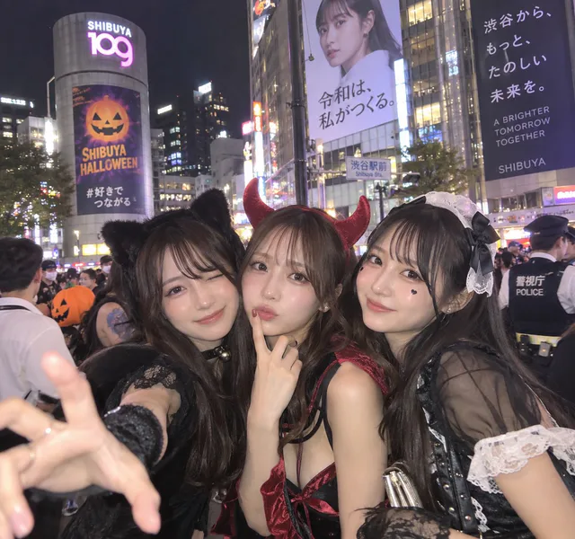 黒い猫耳衣装、赤い悪魔衣装、黒白のメイド衣装を身につけた3人の成人女性が、夜の渋谷で顔を寄せて自撮りするAI生成画像