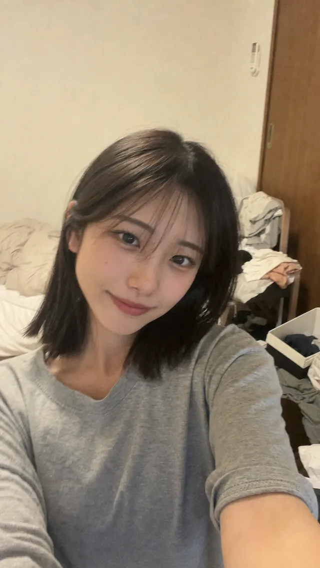 散らかった寝室で、グレーのTシャツを着た可愛い成人の日本人女性が少し手ブレしたスマートフォン自撮りをしているAI生成画像