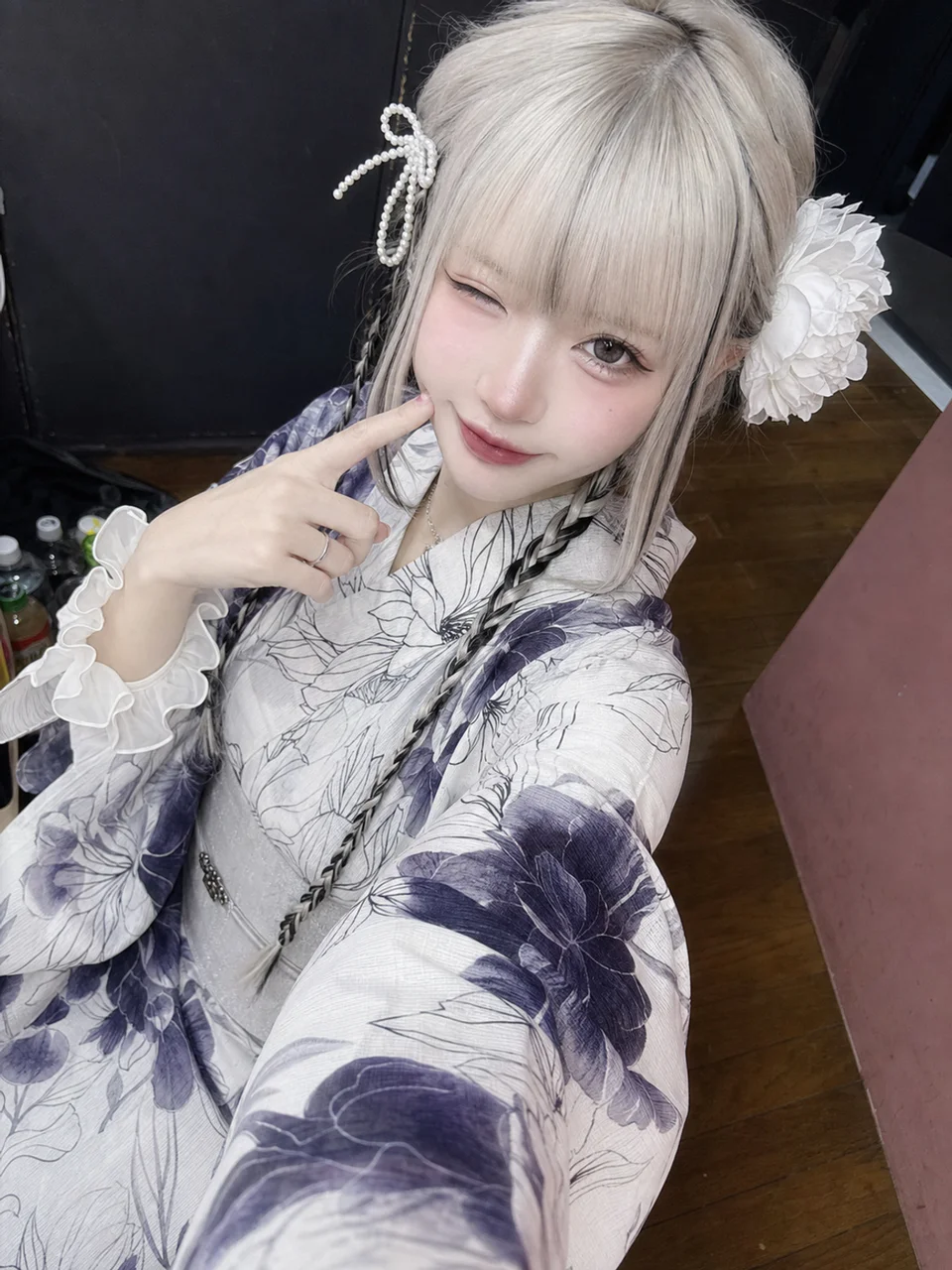 紫の花柄浴衣を着たプラチナブロンドの成人女性が、真珠のリボンと白い花を髪に飾り、頬に指を添えてウィンクする俯瞰セルフィー