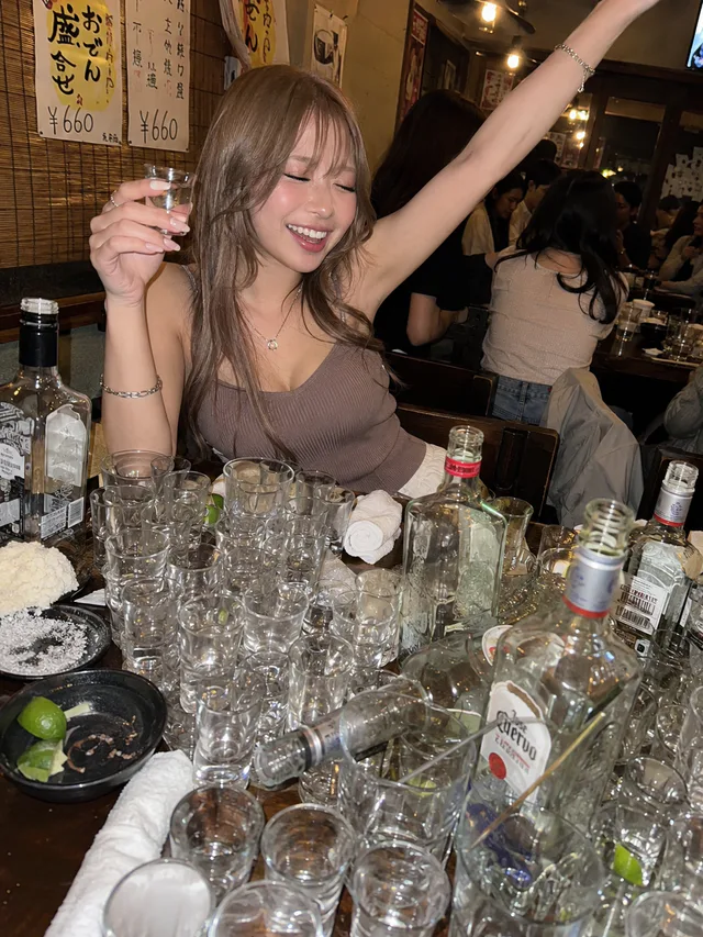 深夜の居酒屋でショットグラスを掲げて笑い、片腕を上げるミルクティー色の長い髪の成人女性と、大量の空グラスやボトルが並ぶテーブル