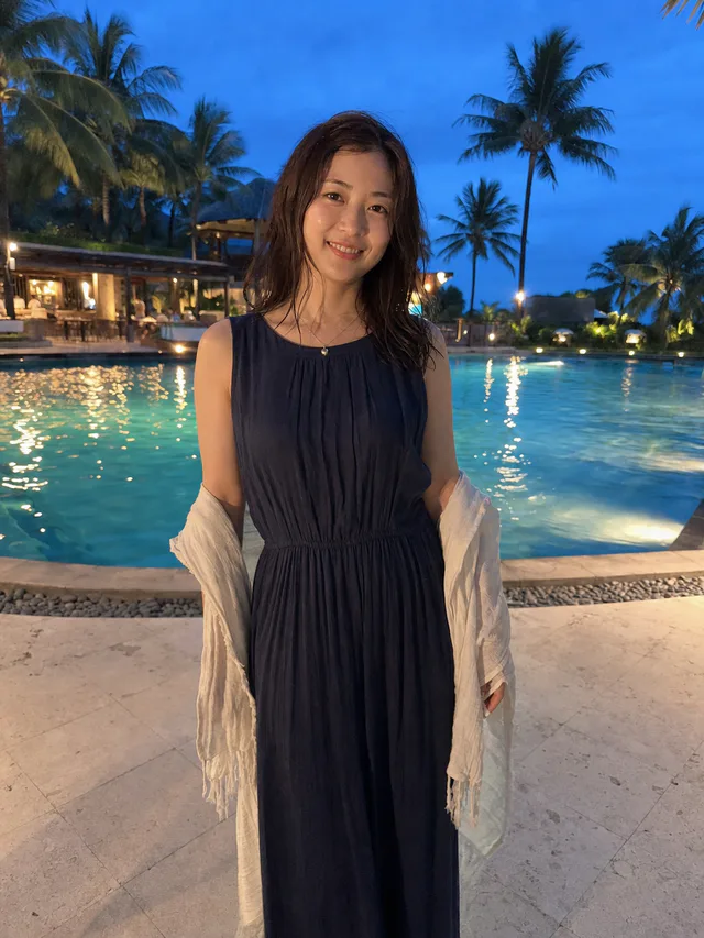 青い夕空とリゾートプールを背景に、ネイビーのロングドレスを着た成人女性が微笑むAI生成のスマホ写真風ポートレート