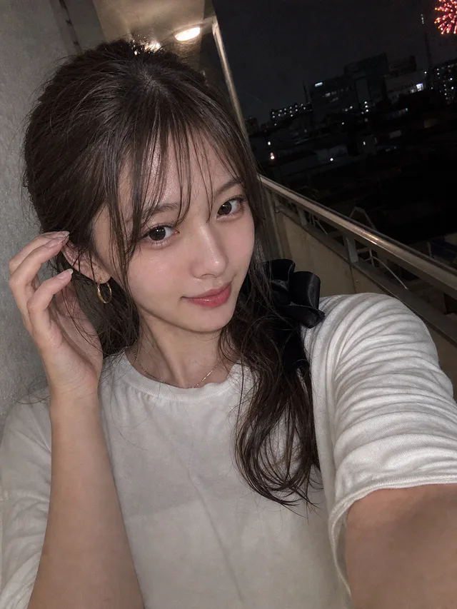 夏の夜のマンション外廊下で、白いTシャツを着た成人女性が遠くの花火と一緒に撮影するAI生成のスマホ自撮り
