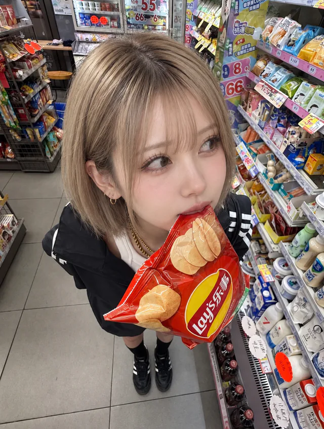 黒いジャケットと黒い靴の白瀬みるが、商品棚の間で赤いお菓子の袋を口にくわえ、横目を向けている俯瞰の一枚。