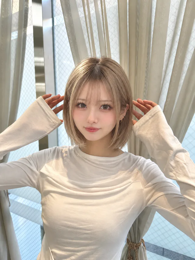 白い長袖トップスの白瀬みるが、カーテンのある窓辺で髪の横に両手を上げ、こちらを見ている。